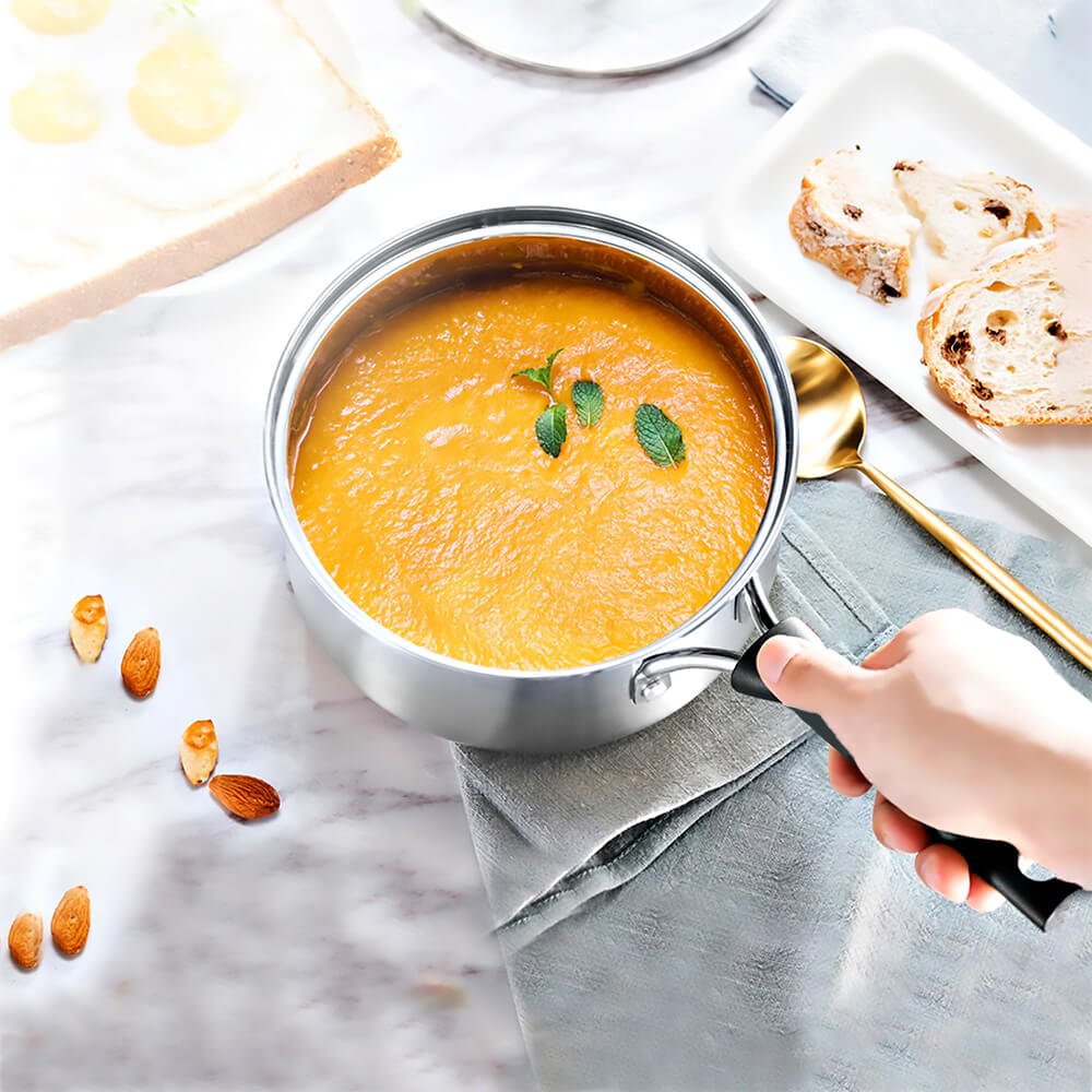 2 quart stainless steel saucepan with lid containing soup, held by a hand with a black handle, placed on a marble surface next to bread and almonds.