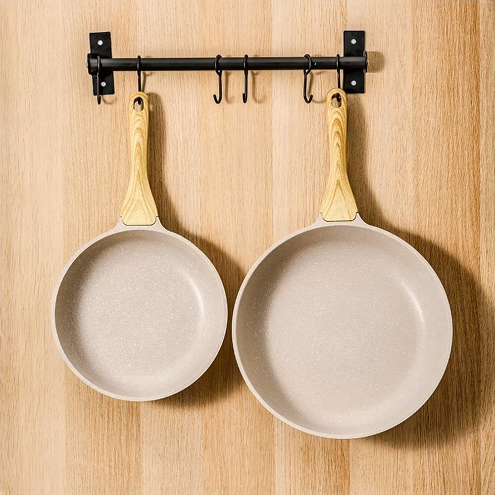 Two aluminum frying pans with wooden handles hanging on a black wall rack against a light wooden kitchen wall.