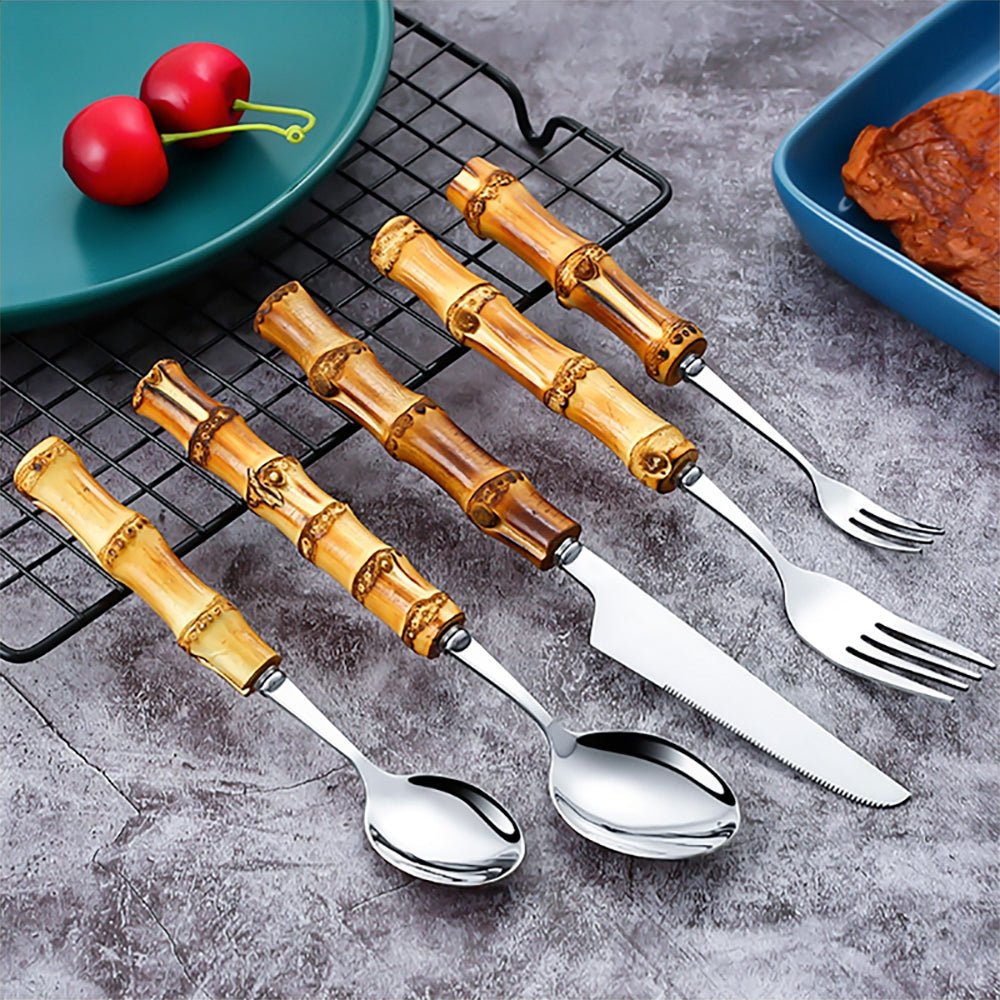 A 5-piece flatware set with bamboo-style handles and stainless steel heads displayed on a textured surface near plates of food.