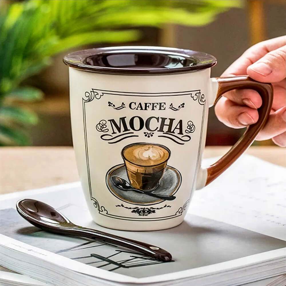 Hand holding the best ceramic mug with lid, featuring a vintage Caffe Mocha design, brown handle, and matching ceramic spoon placed on a magazine.
