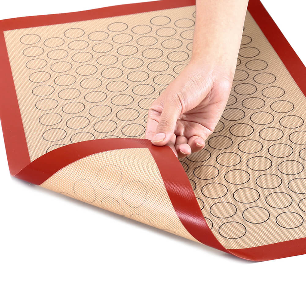 Hand holding flexible best macaron silicone baking mat with circular guides, non-stick surface, and red border for easy lifting.