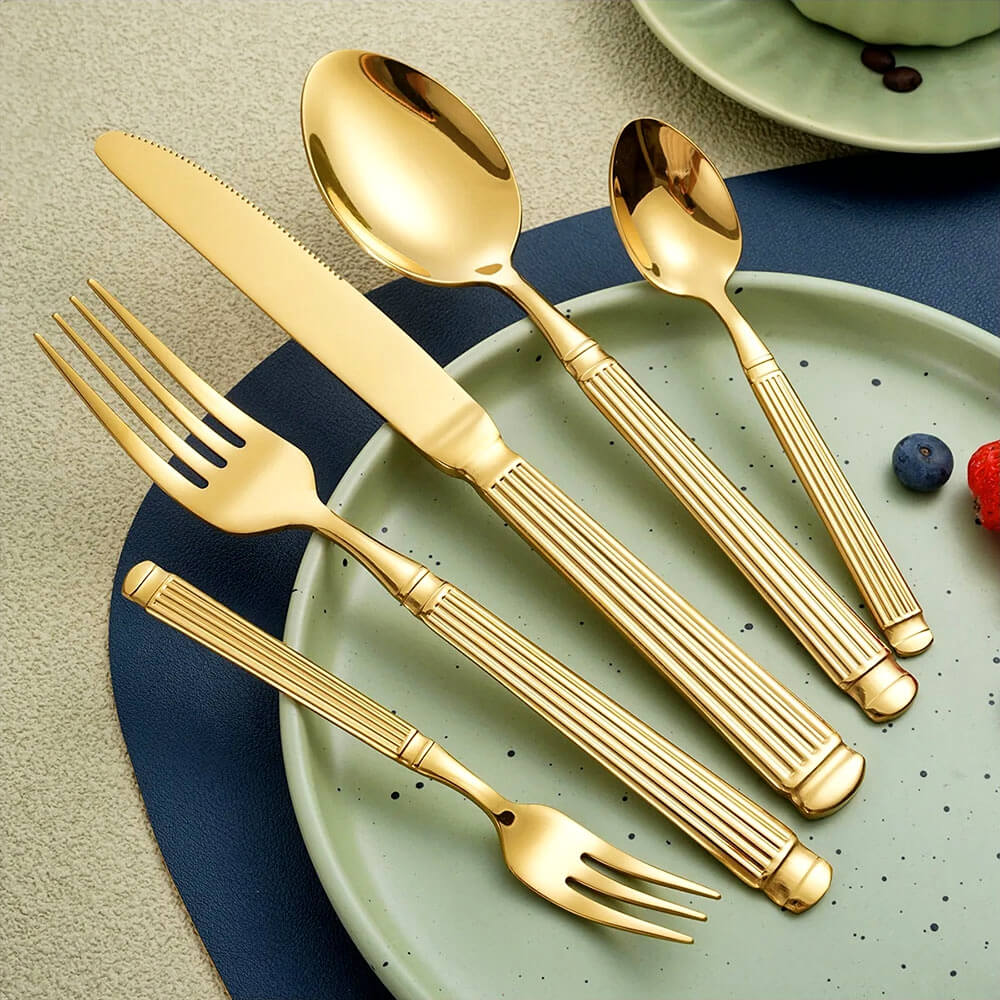 Modern gold flatware set with grooved handles, including knife, forks, and spoons, displayed on a green ceramic plate with a blue placemat.