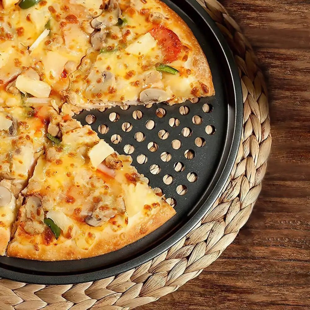 Close-up of a cheese and veggie pizza on a perforated pizza pan, resting on a woven mat atop a wooden surface.