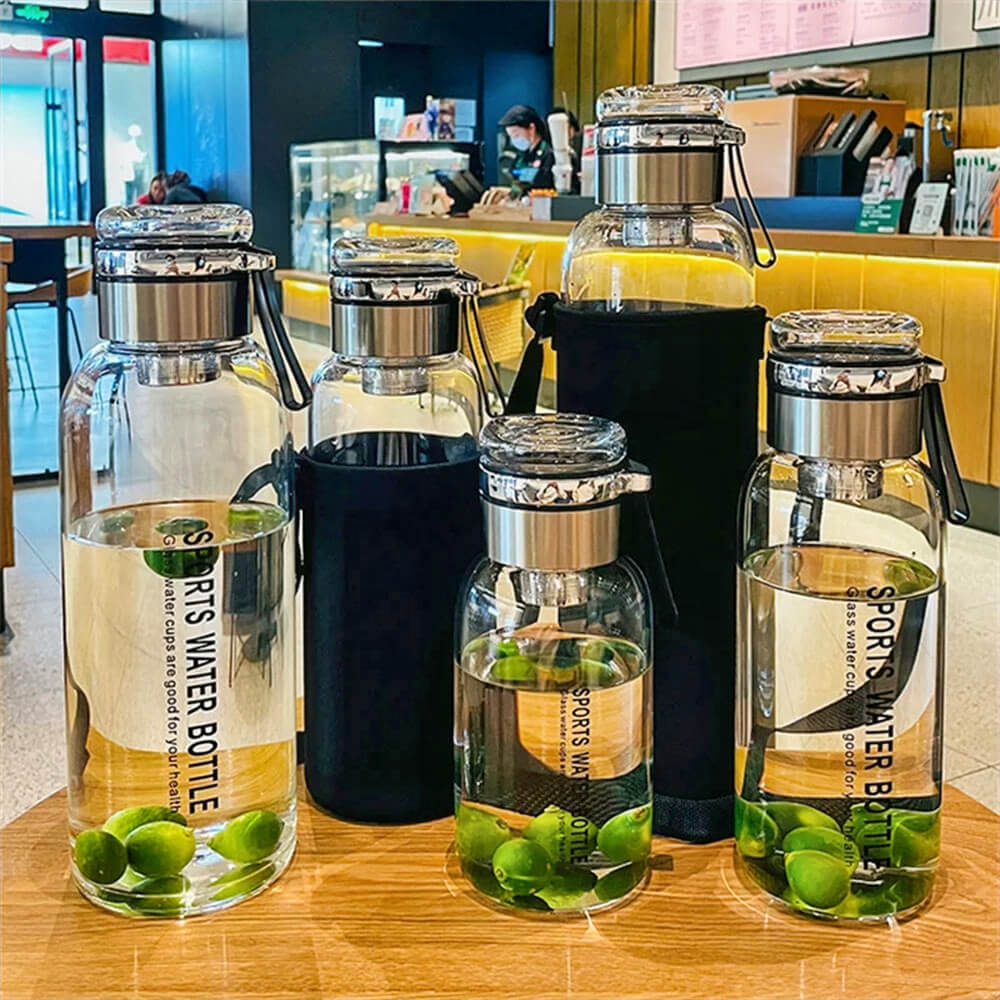 A set of glass sports water bottles in various sizes, featuring stainless steel leakproof lids and neoprene bottle sleeves, displayed on a wooden table. Some bottles contain water with green limes, highlighting their suitability for infused drinks. The background features a modern café setting with warm lighting and a customer service counter.