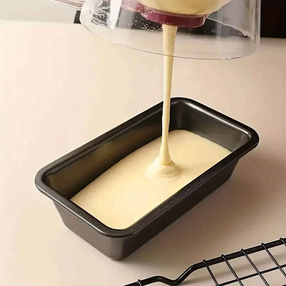 A bread loaf pan carbon steel sits on a light-colored surface as smooth batter is poured into it from a clear mixing container. The pan has a nonstick surface and slightly rounded edges. A black cooling rack is partially visible in the foreground.
