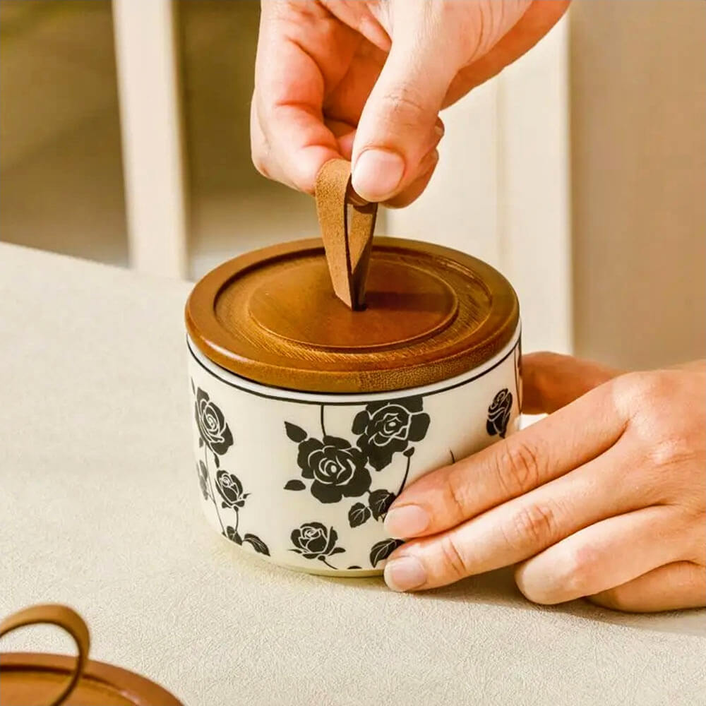 Hand holding a ceramic canister for kitchen with a floral black rose pattern and a wooden lid featuring a leather loop handle, ideal for storing dry goods like sugar, tea, or spices.