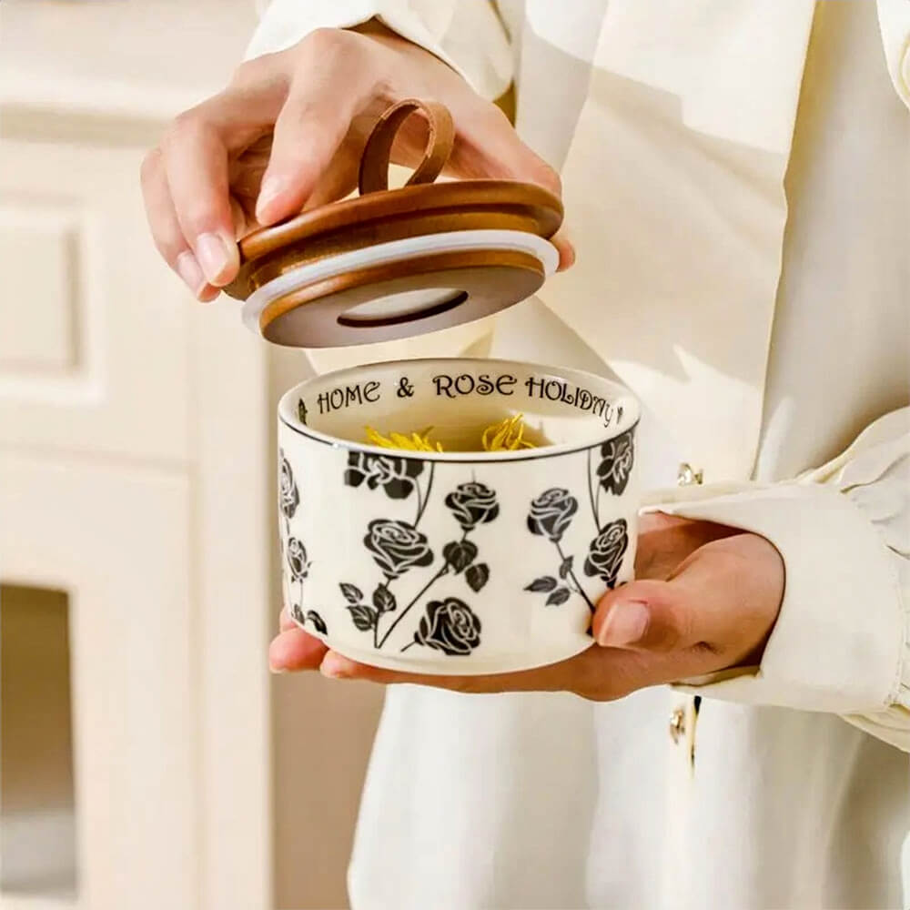 Person holding one of the canisters sets for the kitchen, featuring a ceramic container with a black rose pattern and a wooden lid with a loop handle. The canister is open, showing dried flowers inside, and has HOME & ROSE HOLIDAY, printed on the inner rim.