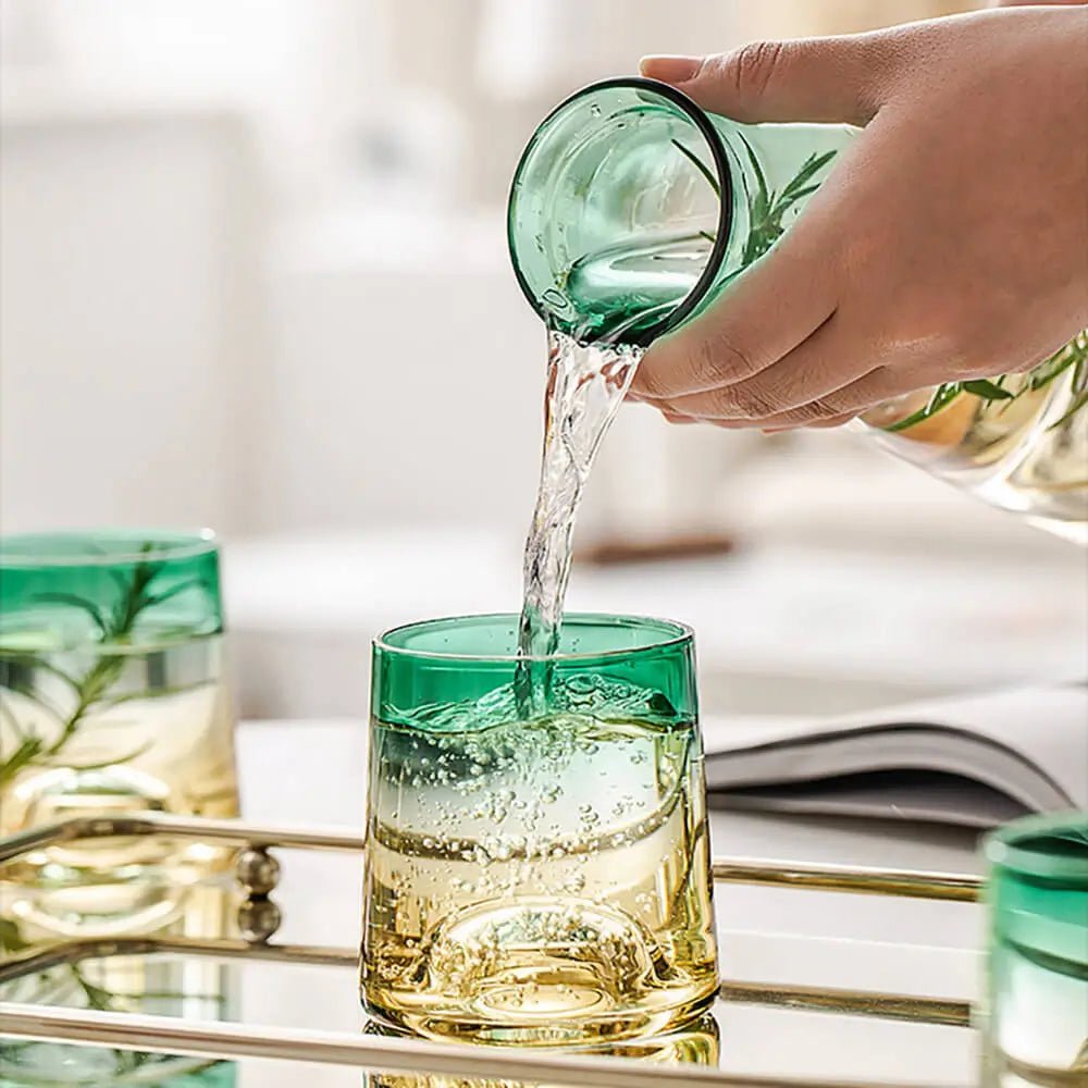 Hand pouring water from a glass carafe into a green and amber glass from a glass carafe set on a mirrored tray.