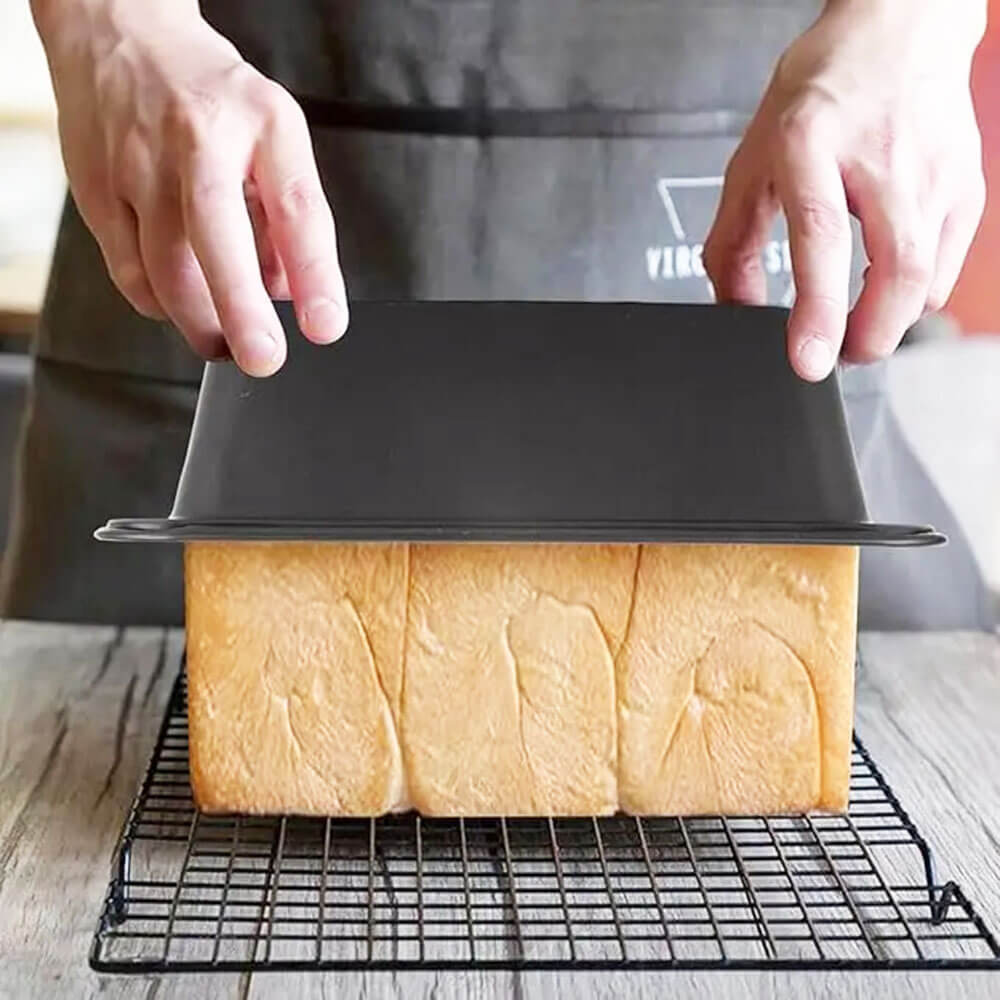 A person wearing a dark apron lifts a carbon steel loaf pan off a freshly baked golden-brown loaf of bread. The bread rests on a black cooling rack placed on a rustic wooden surface. The loaf has a soft texture with defined sections.