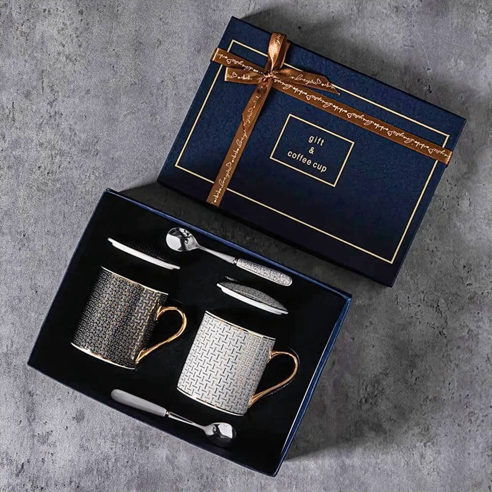 An elegant gift set featuring two ceramic coffee mugs with lids, beautifully arranged in a navy-blue box with gold detailing and a decorative ribbon. Each mug is adorned with a refined geometric pattern and gold-accented handles, accompanied by matching ceramic lids and stainless steel spoons.