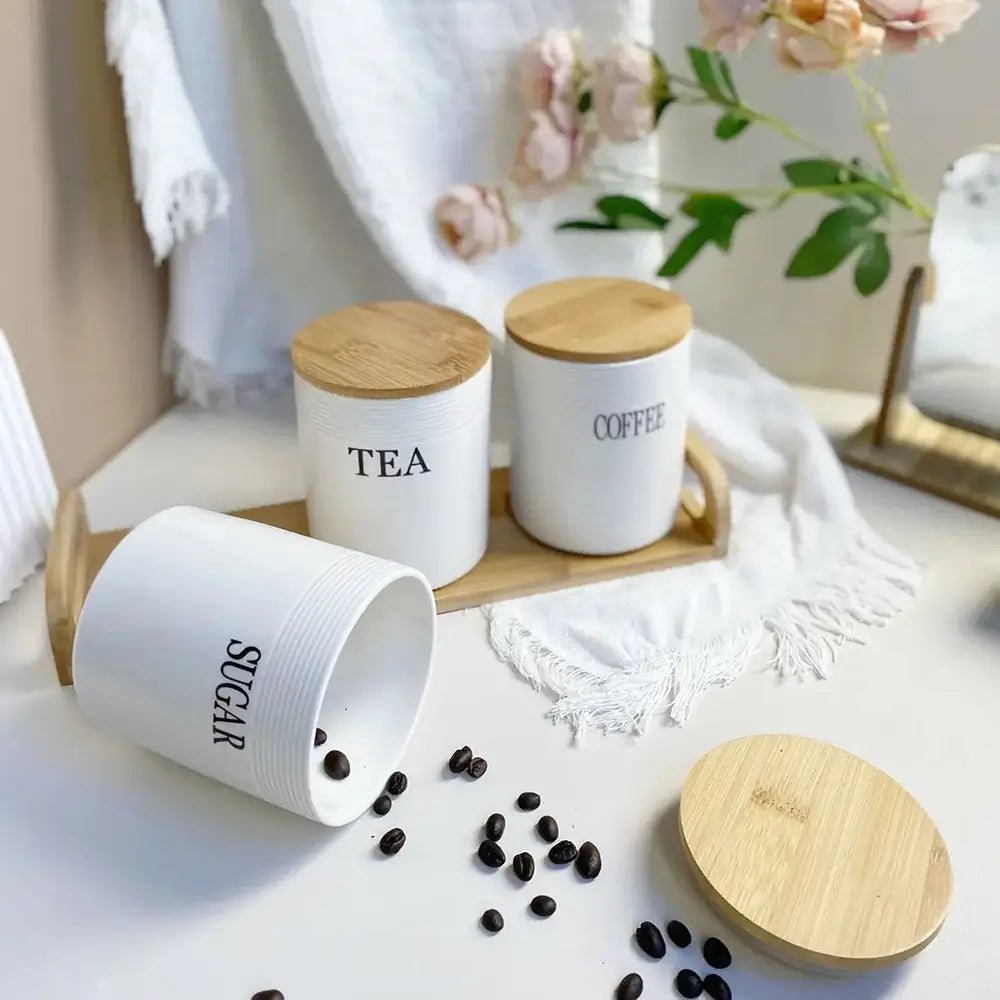 Ceramic tea coffee and sugar canisters with bamboo lids on a wooden tray, one canister tipped over spilling coffee beans.