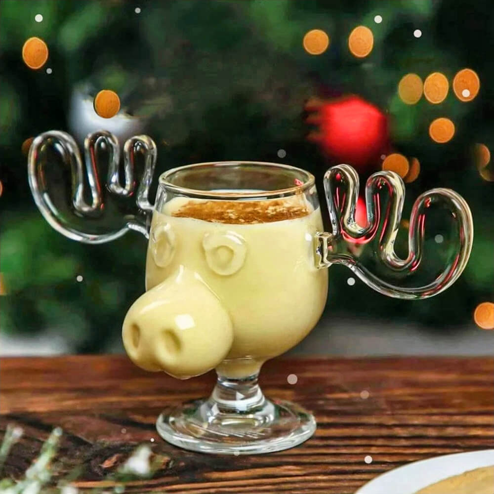 A single glass from the Christmas Vacation Moose Mugs Set of 4 is filled with a creamy beverage, garnished with a sprinkle of cinnamon. The mug features a playful moose design with detailed antlers as handles, set against a festive background with warm holiday lights and a decorated Christmas tree.