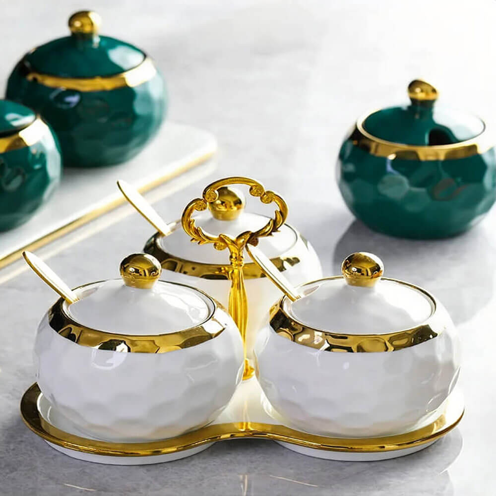 Elegant white and gold condiment containers with lids and spoons, placed on a matching reusable tray with a decorative gold handle. Green containers are visible in the background.