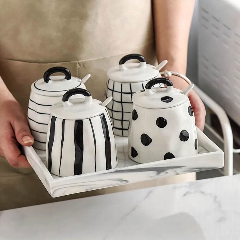Person holding a tray with four ceramic condiment containers with lids, each featuring a black and white design with matching spoons and marble tray.