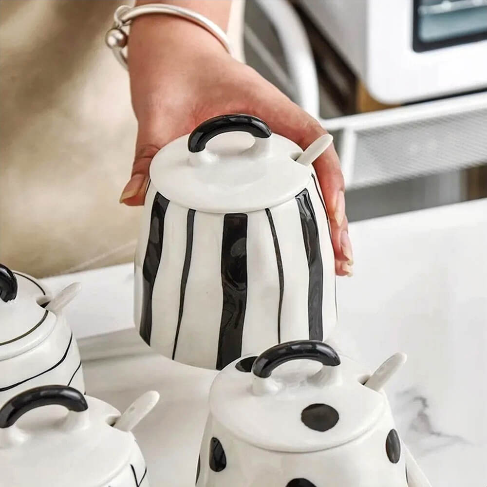 Hand holding one of four ceramic condiment storage containers, featuring a vertical black stripe design with lid and spoon, placed on a marble surface.