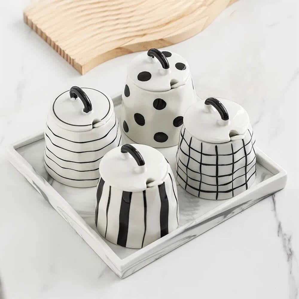 Set of four ceramic condiments container set with black and white patterns, matching lids, and a marble tray, placed on a white countertop.