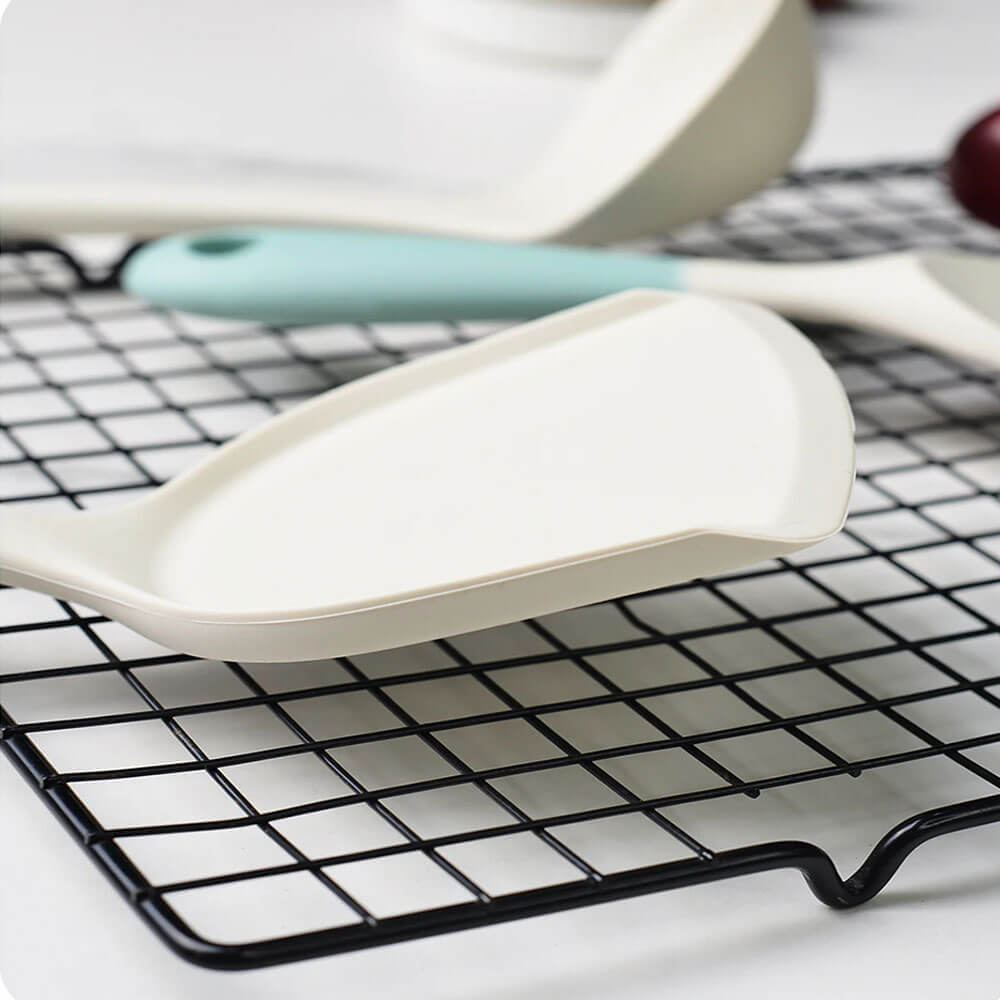 A close-up view of a core kitchen silicone utensil set, featuring a white silicone spatula with raised edges resting on a black cooling rack. In the background, additional utensils with pastel-colored handles are partially visible. The utensils have a modern design with smooth, non-stick surfaces, ideal for cooking and baking.