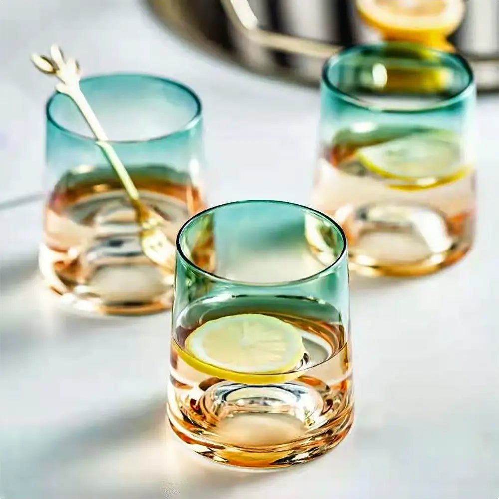 Three gradient green and amber glasses from a crystal carafe and glass set, filled with water and lemon slices, on a white surface.