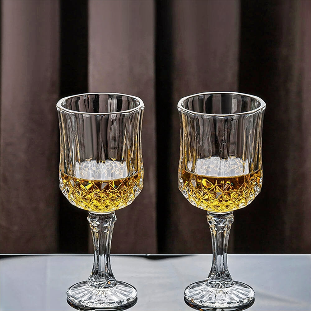 Two crystal glasses with a diamond-cut pattern, filled with amber liquid, placed on a reflective surface against a dark curtain background.