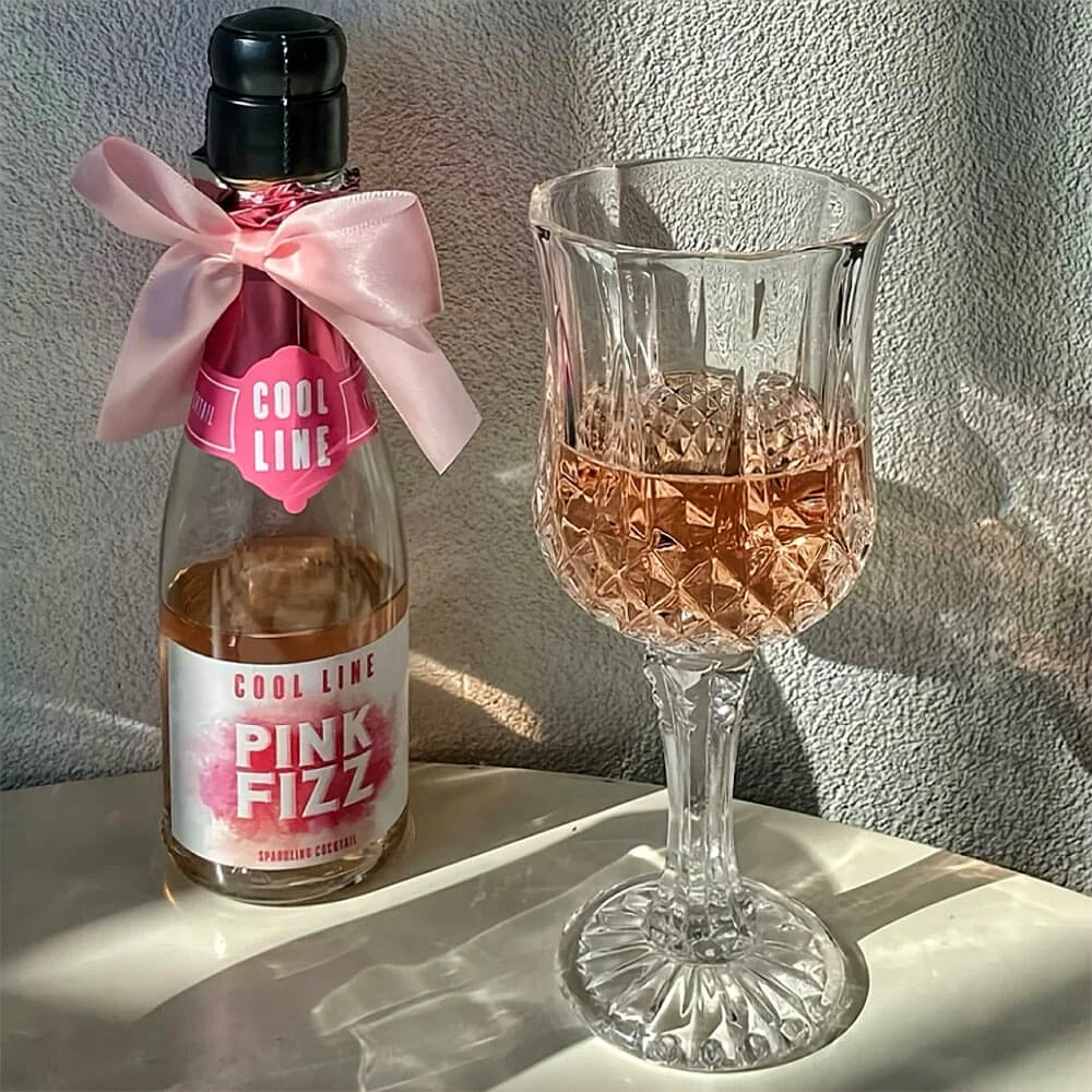 Crystal drinking glass with a diamond-cut pattern filled with pink cocktail and ice, placed beside a bottle of Pink Fizz sparkling cocktail.