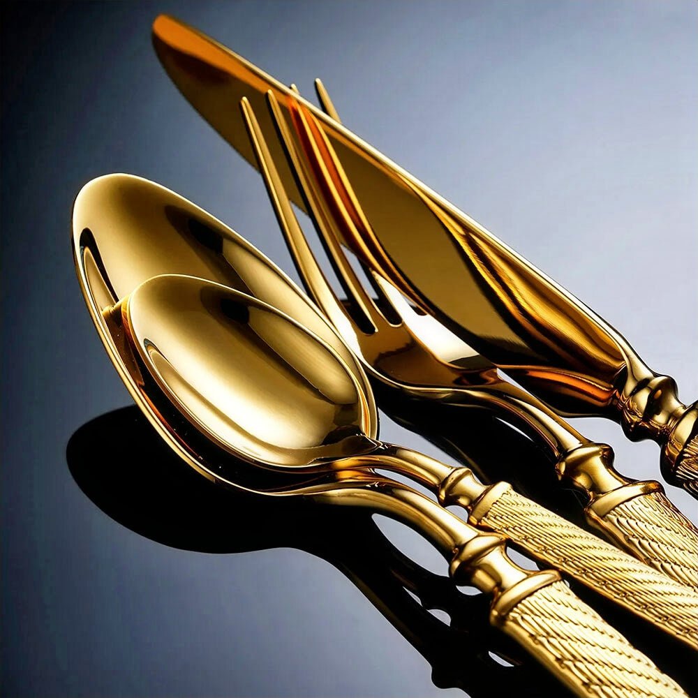 A close-up of a gold cutlery flatware set, including a spoon, fork, and knife. The utensils have a reflective, polished surface with textured handles. The set is arranged on a dark background, highlighting the luxurious gold finish and intricate detailing.
