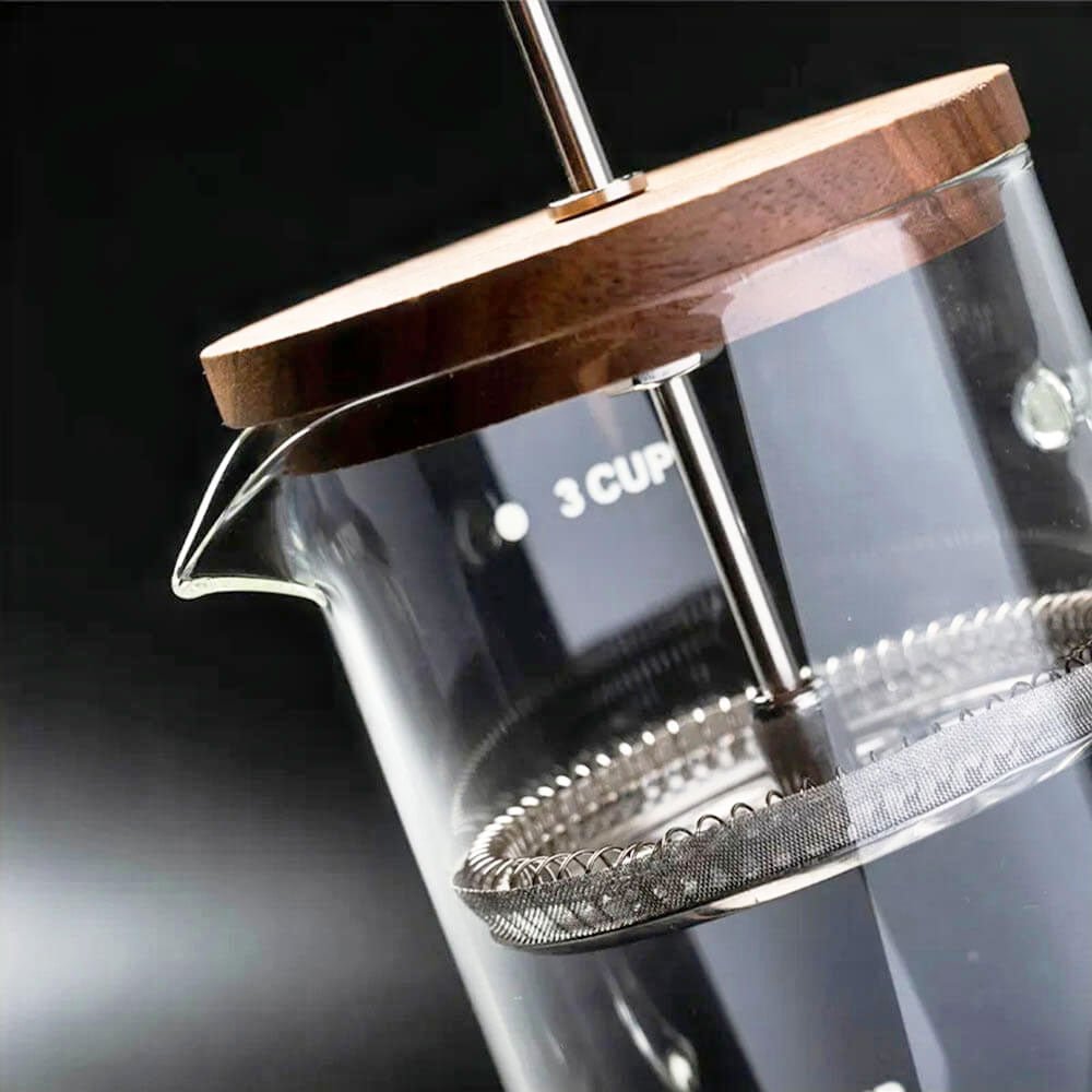 A close-up shot of a glass French press showcases its walnut wood lid, stainless steel plunger, and fine mesh filter inside the transparent glass body. The spout is elegantly curved for easy pouring, and the 3-cup measurement marking is visible on the side. The background is dark, creating a contrast that highlights the sleek design and high-quality materials of the French press