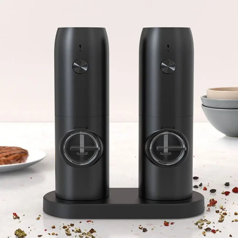 Sleek black electric salt and pepper mill set on stand, with spices scattered on white counter and bowls in background.