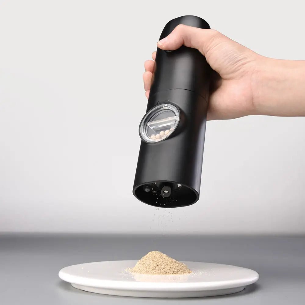 Hand using electric salt and pepper mill to grind seasoning onto a white plate, with peppercorns visible through transparent window.