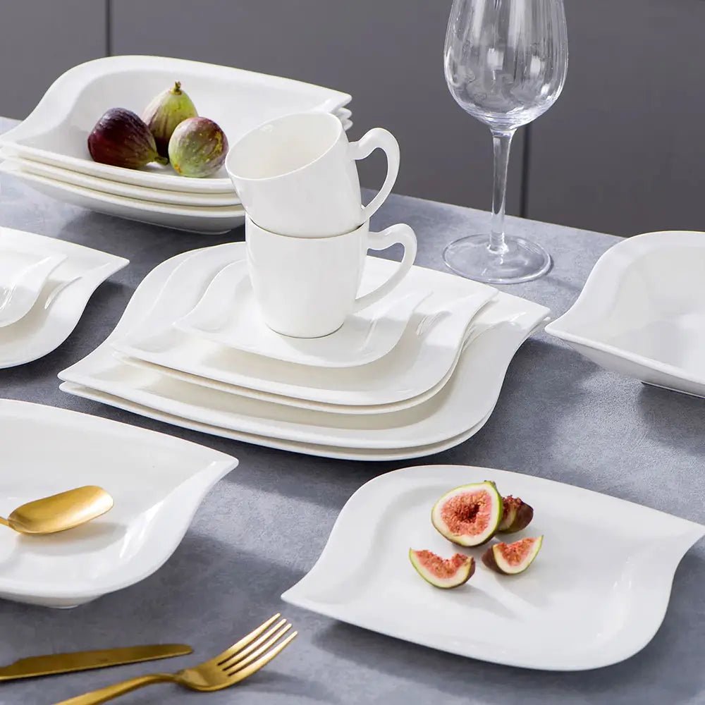 Everyday dinnerware sets with white square and leaf-shaped plates, stacked cups, gold cutlery, figs, and a clear wine glass on a table.