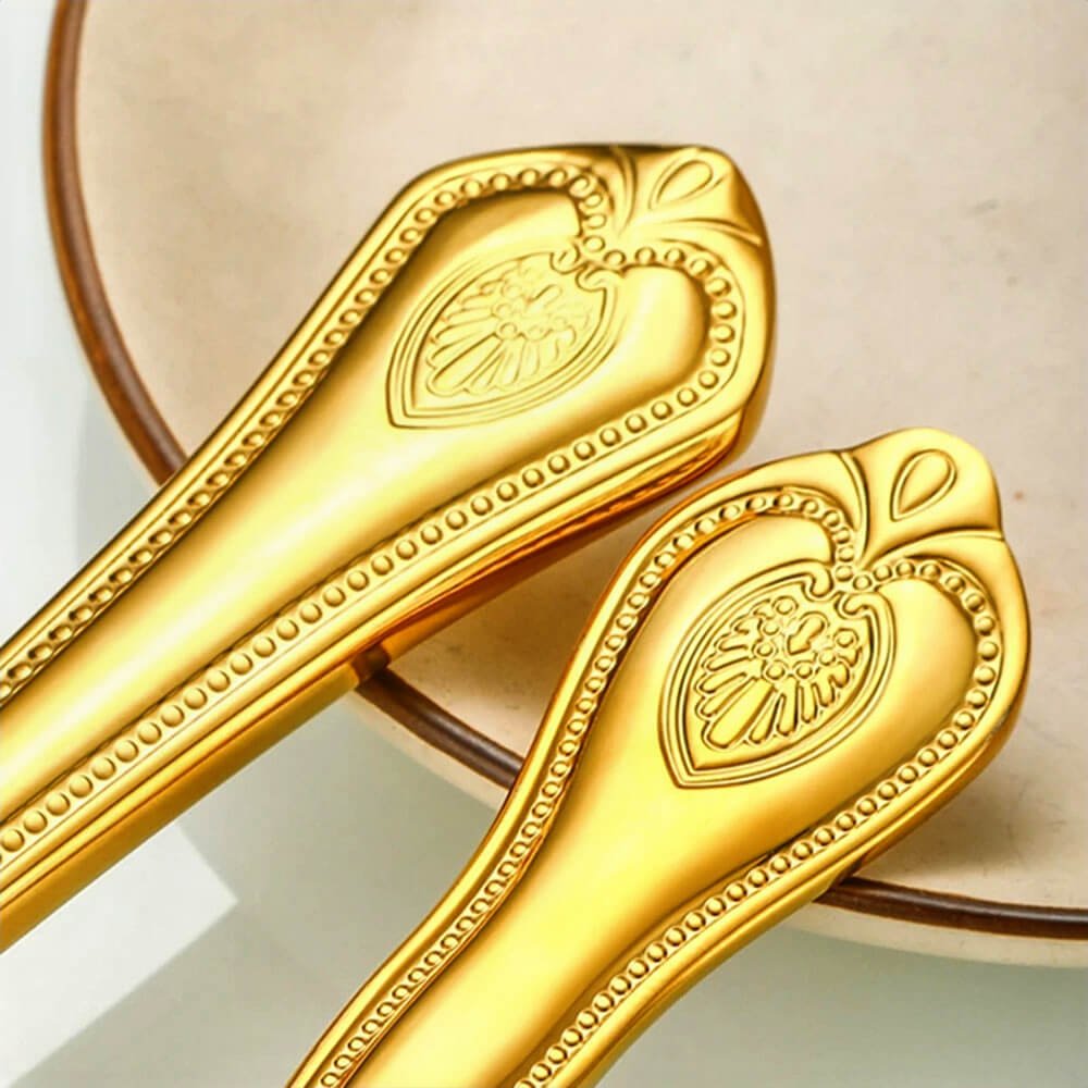 Close-up of two gold flatware handles with ornate embossed detailing, placed beside a ceramic plate.