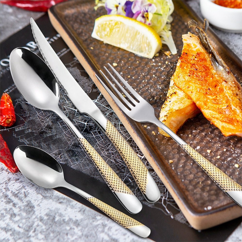 A grilled salmon fillet with lemon and salad is served on a rectangular plate beside a modern flatware set with gold-textured handles.