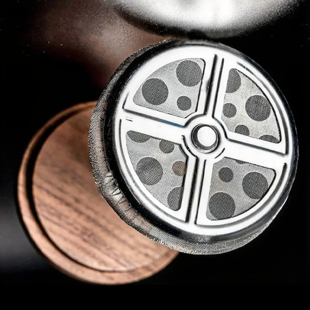 A close-up shot of a glass French press, focusing on the fine stainless steel mesh filter and plunger mechanism. The transparent glass body reveals measurement markings, with "3 CUP" clearly visible. The walnut wood lid sits atop, secured by a central stainless steel rod connected to the plunger.