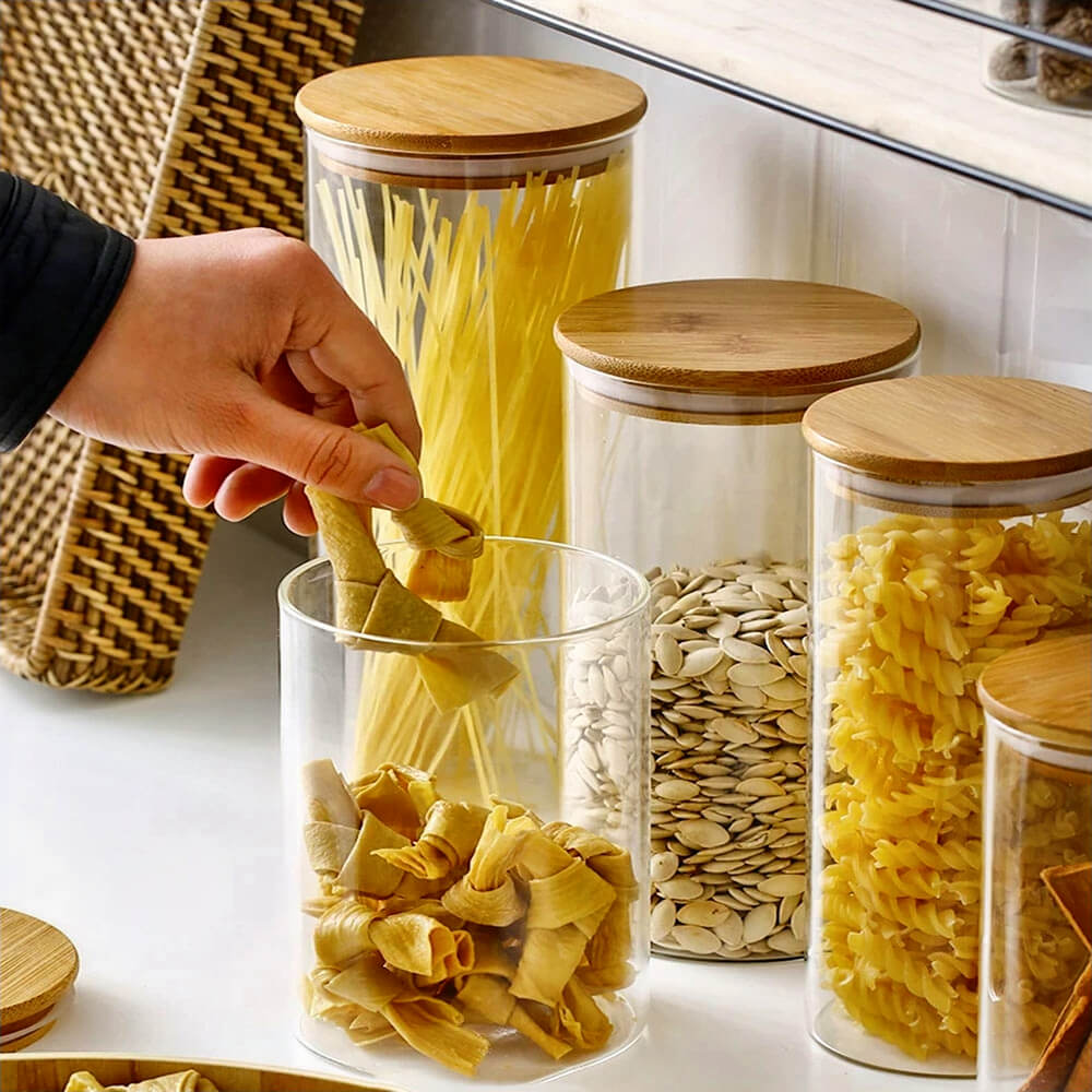 A set of glass and bamboo storage jars filled with various dry goods including pasta and seeds. The jars feature clear cylindrical glass bodies and flat bamboo lids with silicone seals. One jar is open, with a hand reaching in to grab dried pasta.