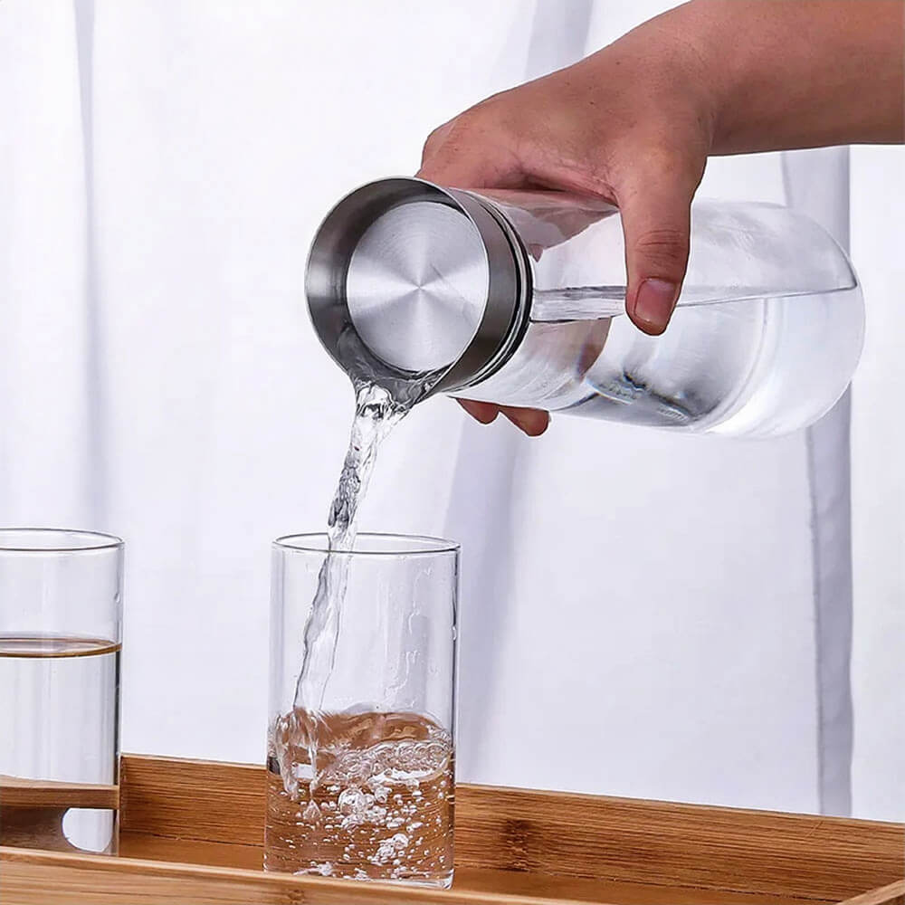 A hand pours water from a glass bottle with a stainless steel leakproof lid into a clear glass. The bottle has a rounded body and wide mouth, ideal for serving glass bottle water at home or in a minimalist kitchen setting.