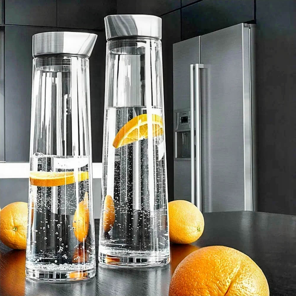 Two tall glass bottles for drinks with stainless steel leakproof lids are filled with sparkling water and orange slices. Set on a dark kitchen counter beside fresh oranges, the bottles have a clear, straight-sided design suited for serving cold beverages.