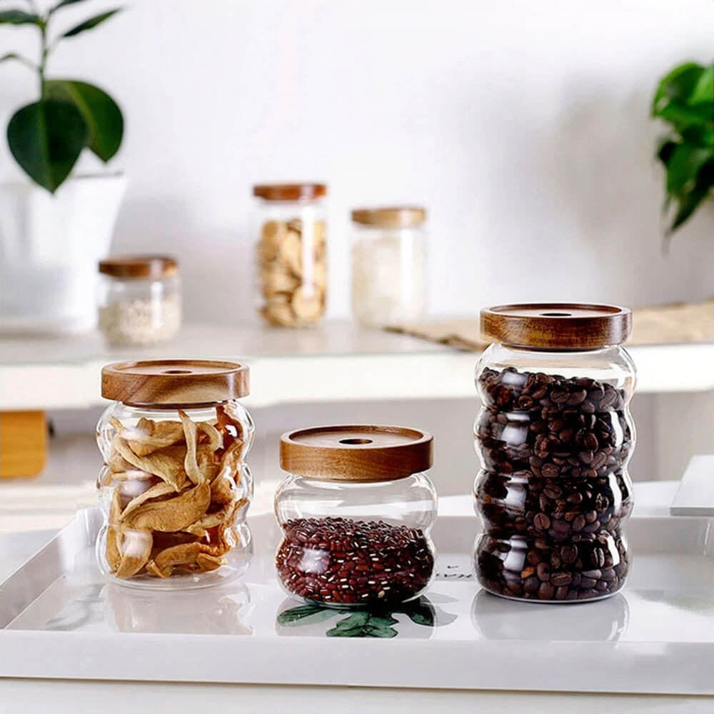 Three ripple-textured glass canisters with natural wooden lids are displayed on a tray, each filled with dried fruit, red beans, and coffee beans. Additional jars are arranged on a shelf in the background, creating a clean and organized kitchen setting.