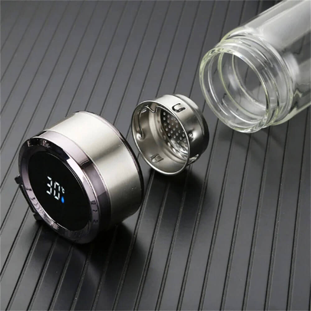 A disassembled glass coffee travel tumbler placed on a ridged black surface, showing the main glass body, a stainless steel infuser, and a digital temperature display lid indicating 30°C. The components highlight the tumbler&