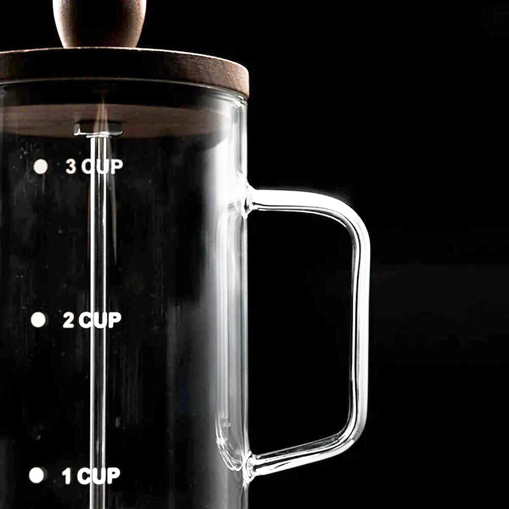 A close-up shot of a glass French press with no plastic, highlighting its sleek, transparent borosilicate glass body and sturdy glass handle. The measurement markings for 1, 2, and 3 cups are visible on the side, ensuring precise brewing. The walnut wood lid is partially visible at the top, adding a natural touch to the minimalist design.
