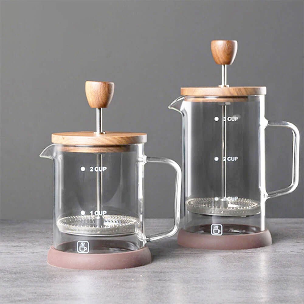 Two glass French presses of different sizes are displayed side by side on a textured gray surface against a neutral gray background. Both feature a clear borosilicate glass body, walnut wood lid, and stainless steel plunger with a fine mesh filter for smooth coffee extraction. The smaller press has a 2-cup capacity, while the larger one holds 3 cups, as indicated by the white measurement markings on the glass.