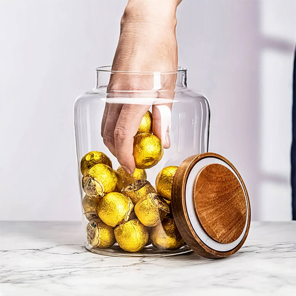 A clear glass jar with wood lid sits on a marble countertop, filled with gold-wrapped chocolates. The airtight wooden lid with a silicone seal is placed beside the jar, showcasing its secure closure. A person&