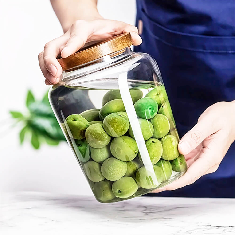 A person wearing a navy apron is holding a clear glass jar with a wooden lid. The jar is filled with green olives submerged in liquid. One hand supports the bottom of the jar while the other secures the lid. The background features a blurred green plant, and the scene is set on a marble countertop. The lighting is bright, emphasizing the transparency of the glass and the freshness of the olives.