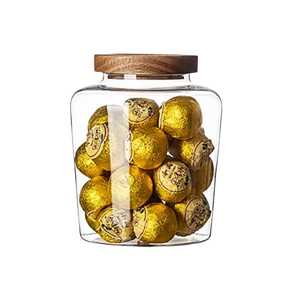 A transparent glass jar with a wooden lid is filled with gold-wrapped chocolates. The jar has a modern, square-shaped design with smooth edges, and the wooden lid features a secure fit, likely with an airtight seal. The chocolates inside are stacked, showcasing the clear visibility of the jar’s contents. The background is plain white, emphasizing the jar&