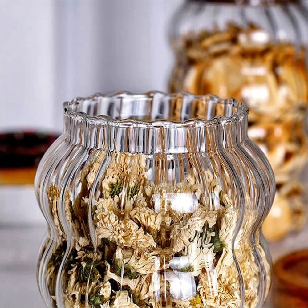 A ripple-textured glass canister filled with dried flowers is shown in the foreground. A second canister with dried food is visible in the blurred background.