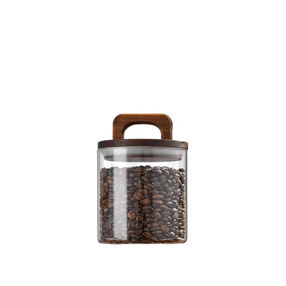 A transparent glass pantry jar filled with coffee beans, featuring a dark wooden lid with a built-in handle, set against a plain white background.