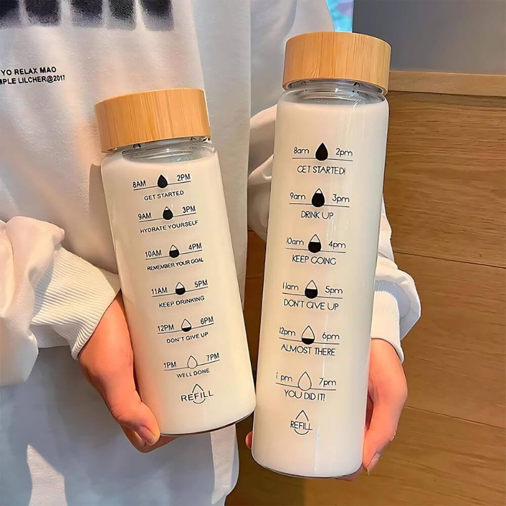 Two glass reusable water bottles with bamboo lids held by a person, featuring time markers from 8 AM to 7 PM and motivational phrases such as GET STARTED, KEEP DRINKING, and YOU DID IT. Designed to encourage consistent hydration throughout the day.