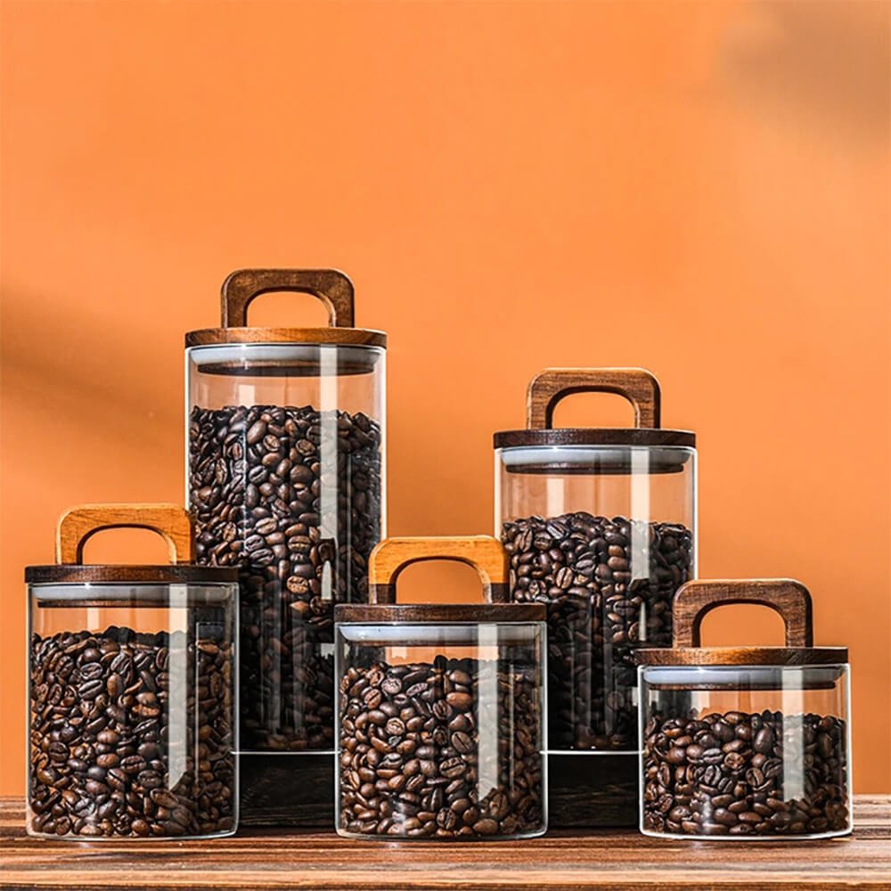 A set of transparent glass storage jars for pantry, filled with coffee beans, arranged on a wooden surface against an orange background. Each jar has a cylindrical shape and features a dark wooden lid with a built-in handle. The jars vary in size, creating a visually appealing display while providing functional storage for pantry essentials.