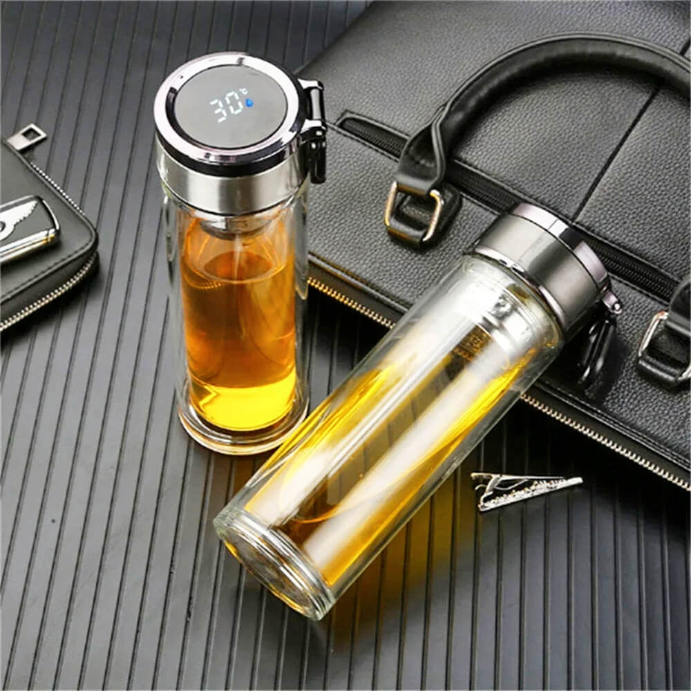 Two stylish glass travel coffee tumblers filled with tea or coffee are placed on a ribbed black surface beside a black leather bag and wallet. One tumbler stands upright displaying a digital temperature of 30°C on the lid, while the other lies flat, showcasing its sleek, double-walled design and built-in carabiner clip for convenience.