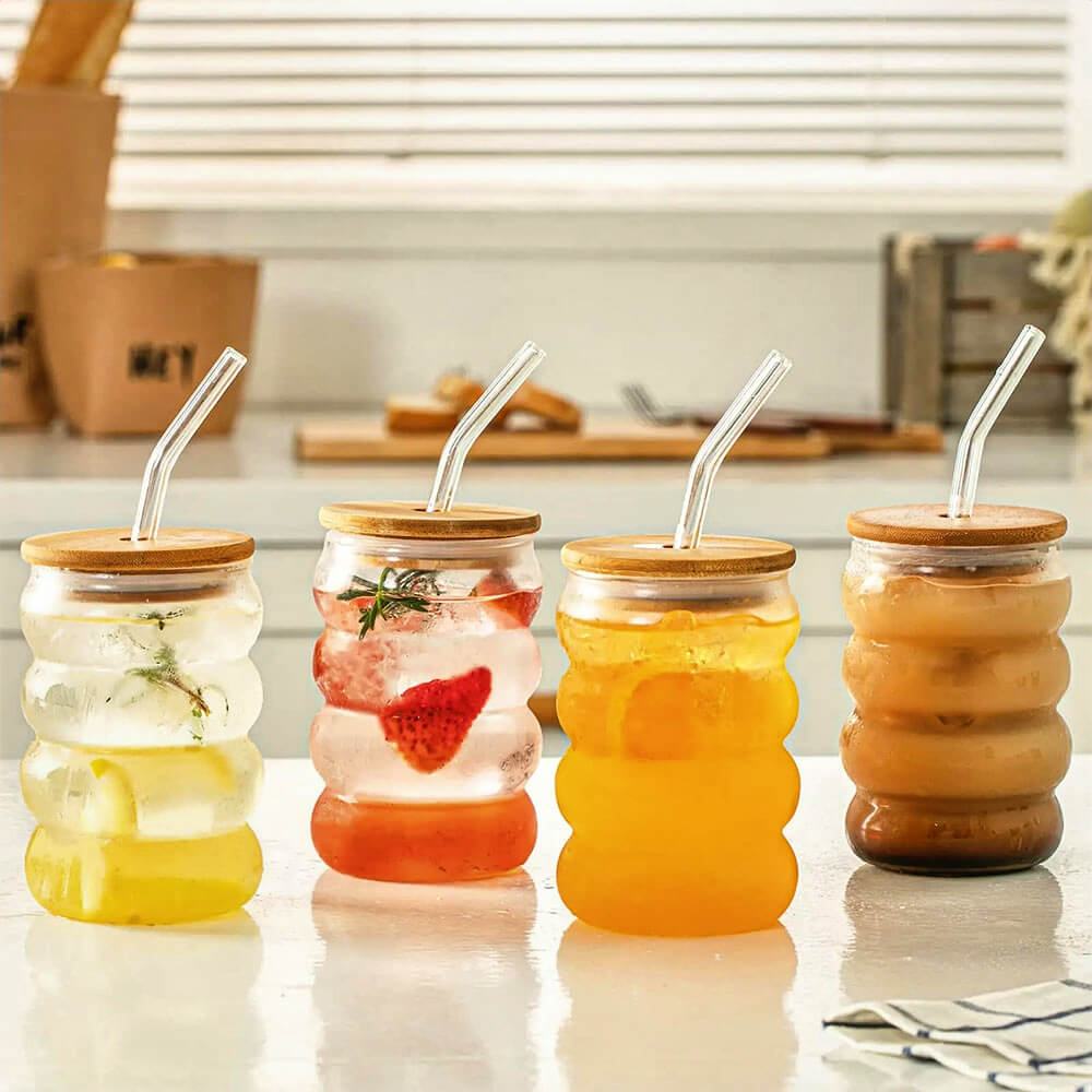 Four wavy glass tumbler with glass straw options, each with a bamboo lid, filled with various beverages including infused water, fruit drinks, and iced coffee, displayed on a bright kitchen counter.