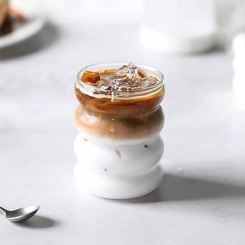 A wavy glass tumbler with lid, filled with iced coffee and milk, served without the lid on, placed on a light countertop with a spoon nearby.
