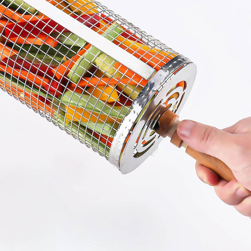 A BBQ grill basket filled with sliced vegetables, held by a person gripping its wooden handle. The stainless steel mesh design allows even cooking while keeping the food contained.